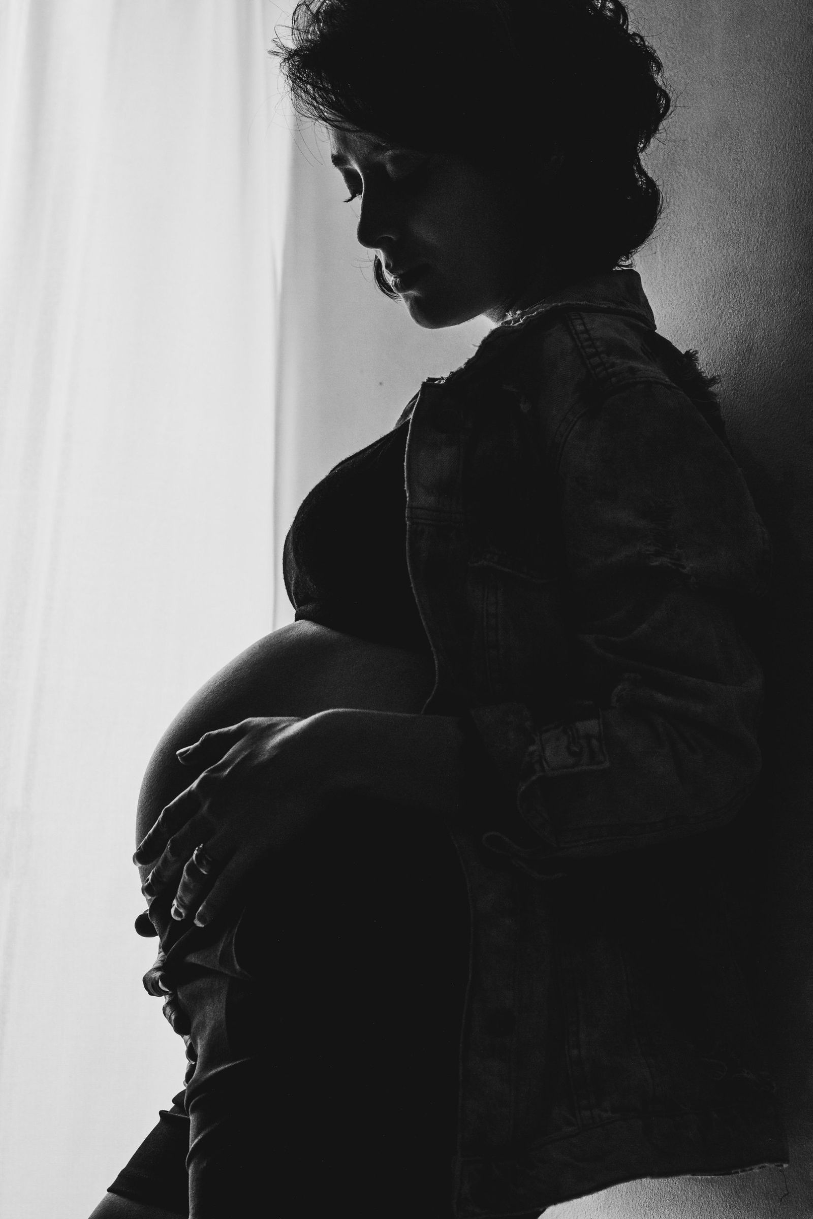 Black and white silhouette of a pregnant woman in a denim jacket, looking down at her bump beside a bright window.
